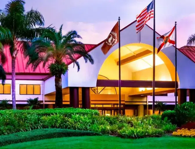Entrance of Sheraton Suites Orlando International Airport Hotel, featuring a grand archway, three flags, lush landscaping, and evening lighting.