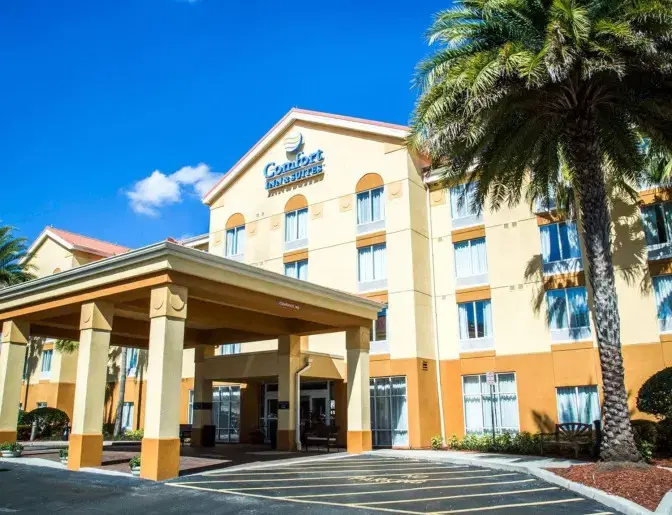 Exterior view of Comfort Inn & Suites Sanford hotel with a covered entrance, palm trees, and a well-maintained landscape under a clear blue sky.