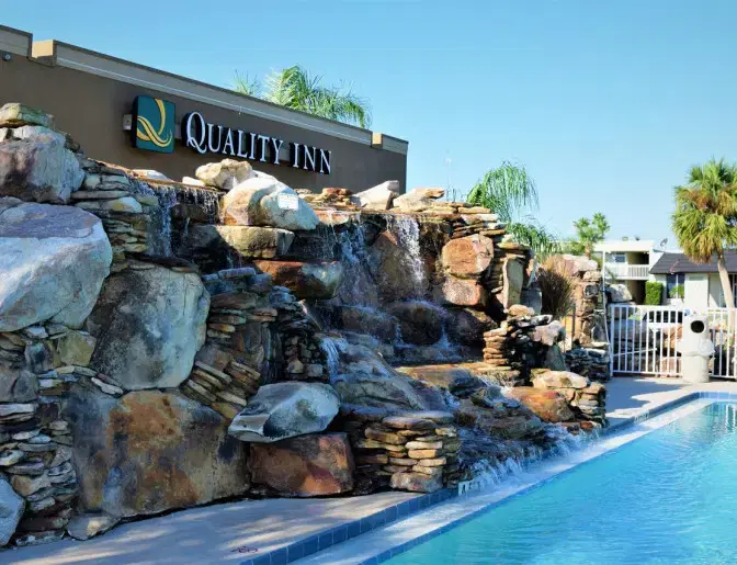 Outdoor view of Quality Inn Titusville Hotel showcasing a rock waterfall and pool area with palm trees in the background.