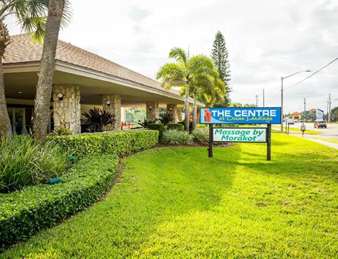Entrance to The Centre at Ocean Landings with a well-maintained garden, palm trees, and a sign along a roadside.