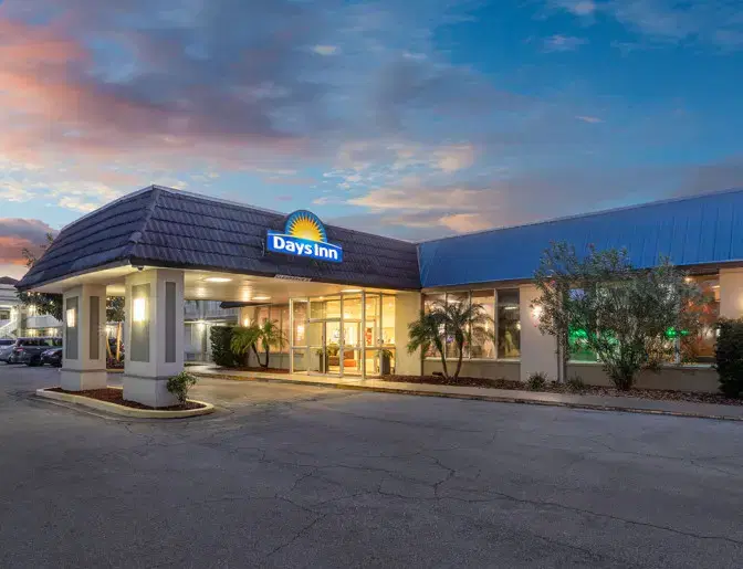 Exterior view of a Days Inn Titusville hotel at dusk, featuring a well-lit entrance, palm trees, and a parking lot with several cars.