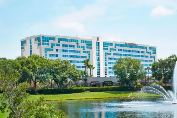 Exterior view of Renaissance Orlando Airport Hotel featuring a modern multi-story building with geometric blue-tinted windows, surrounded by palm trees, a grassy lawn, and a fountain in a reflective pond.