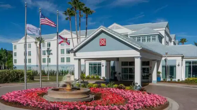 Exterior view of Hilton Garden Inn Orlando Airport Hotel during the day, featuring the main entrance with a water fountain surrounded by pink flowers and palm trees