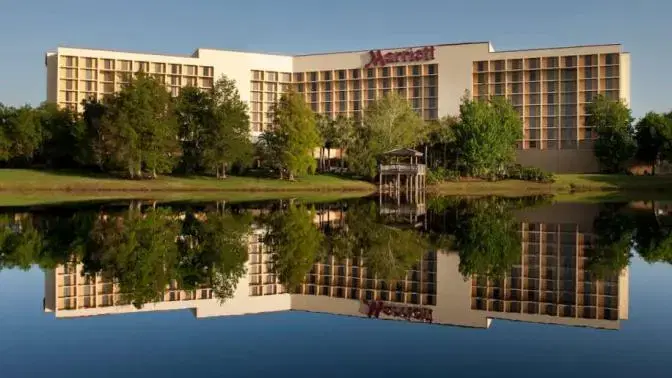 Exterior view of the Marriott Orlando Airport Lakeside Hotel featuring modern architecture surrounded by lush greenery, reflected in a calm lake under a clear blue sky.