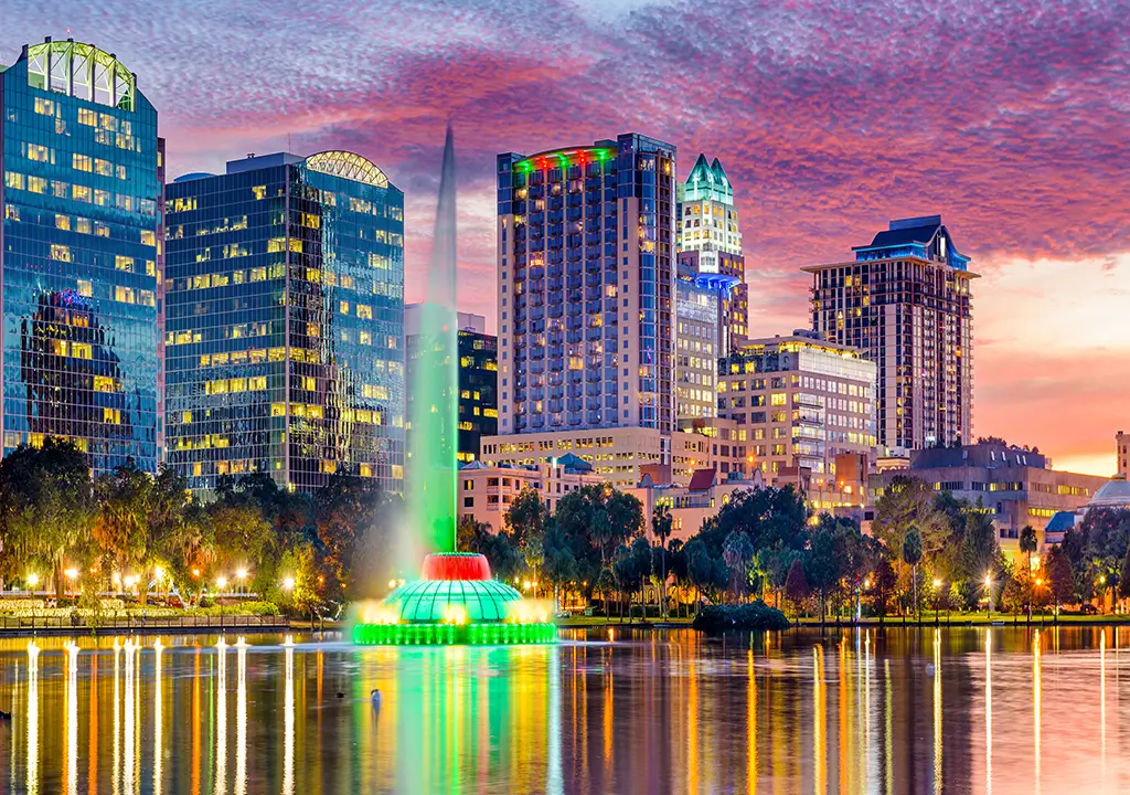 orlando florida skyline with sunset behind buildings and buildings reflected in lake with fountain lit green