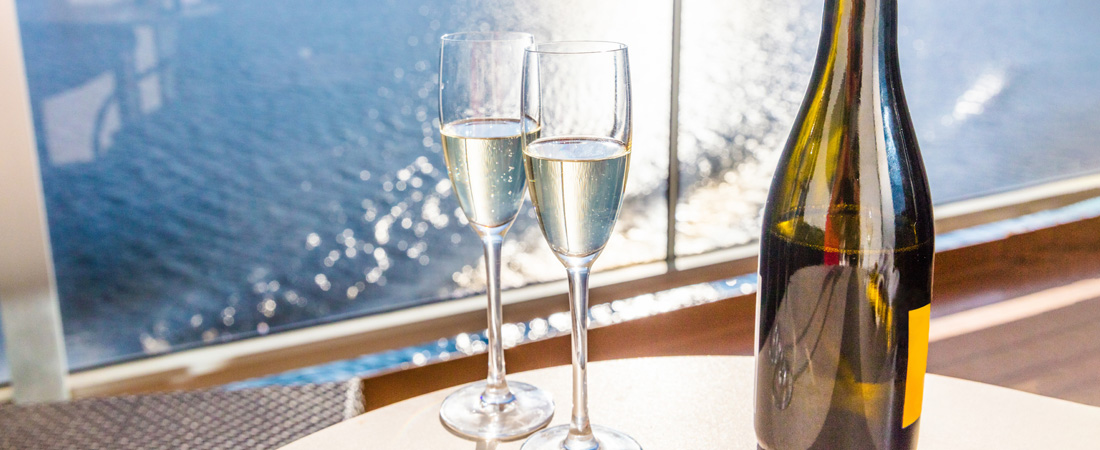 two wine glass flutes and a bottle of wine on a coffee table by the cruise balcony with the sea in the background. one of the cruise mistakes to avoid is leaving a wine bottle back at home