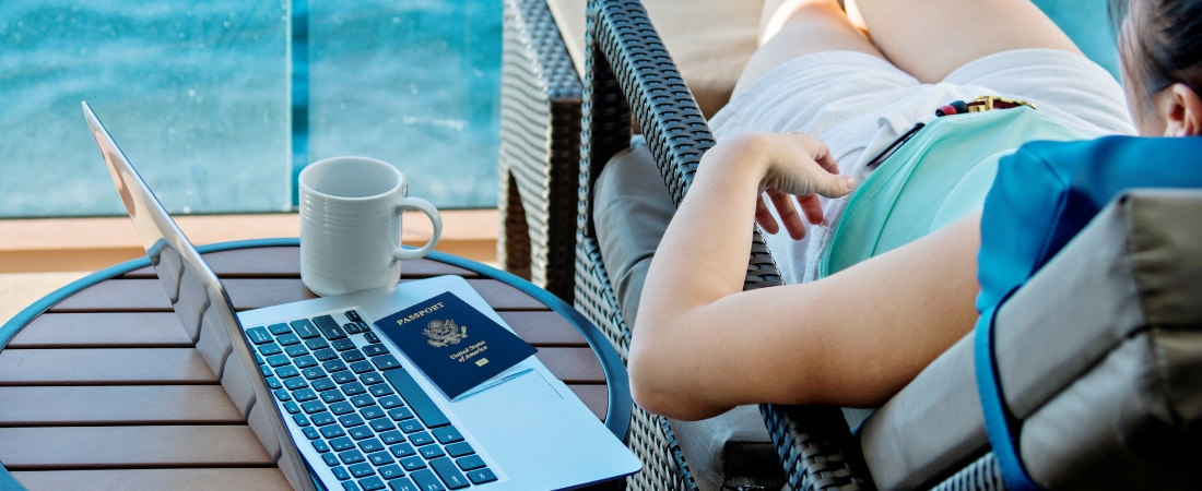 woman lounging on cruise deck looking out at sea, sitting next to coffee table with coffee mug, laptop, and U.S working passport on top of the laptop. one of the cruise mistakes to avoid is not carrying a working passport