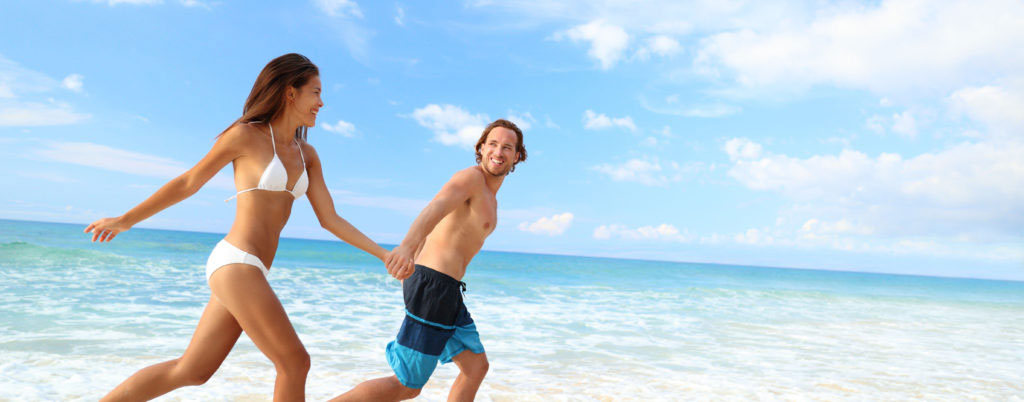 Happy beach couple vacation getaway. Young people in bikini and swimwear running holding hands together having fun on tropical beach paradise. Perfect blue ocean water and white sand.