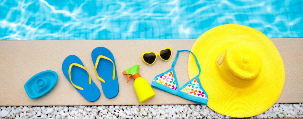 Swimming pool accessories flat lay. Top view of beach items on pool deck. Flip flops bikini and hat sun glasses. Water toys. Summer vacation in tropical resort. Copy space. Colorful beach wear.