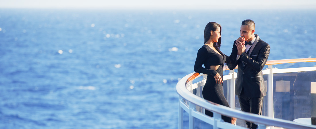 Couple in black formal outfits: man in a black tuxedo is kissing the hand of a woman in a black dress on a cruise balcony top deck, blue ocean in the background