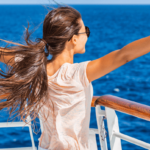 A woman in a light colored shirt standing at the edge of a ship dock with her arms spread open towards the ocean in front of her