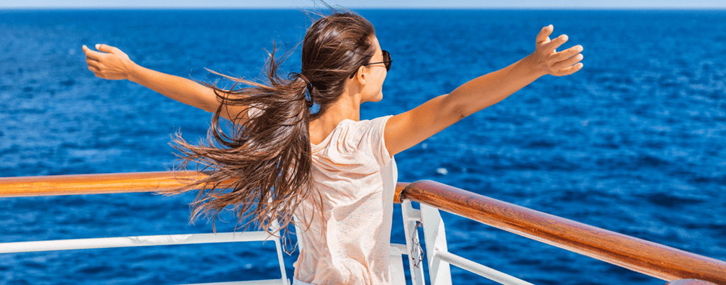 A woman in a light colored shirt standing at the edge of a ship dock with her arms spread open towards the ocean in front of her