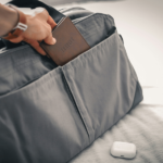A person places a brown travel wallet labeled “TRAVELS” into the front pocket of a gray travel bag resting on a bed, with a pair of white wireless earbuds beside it.