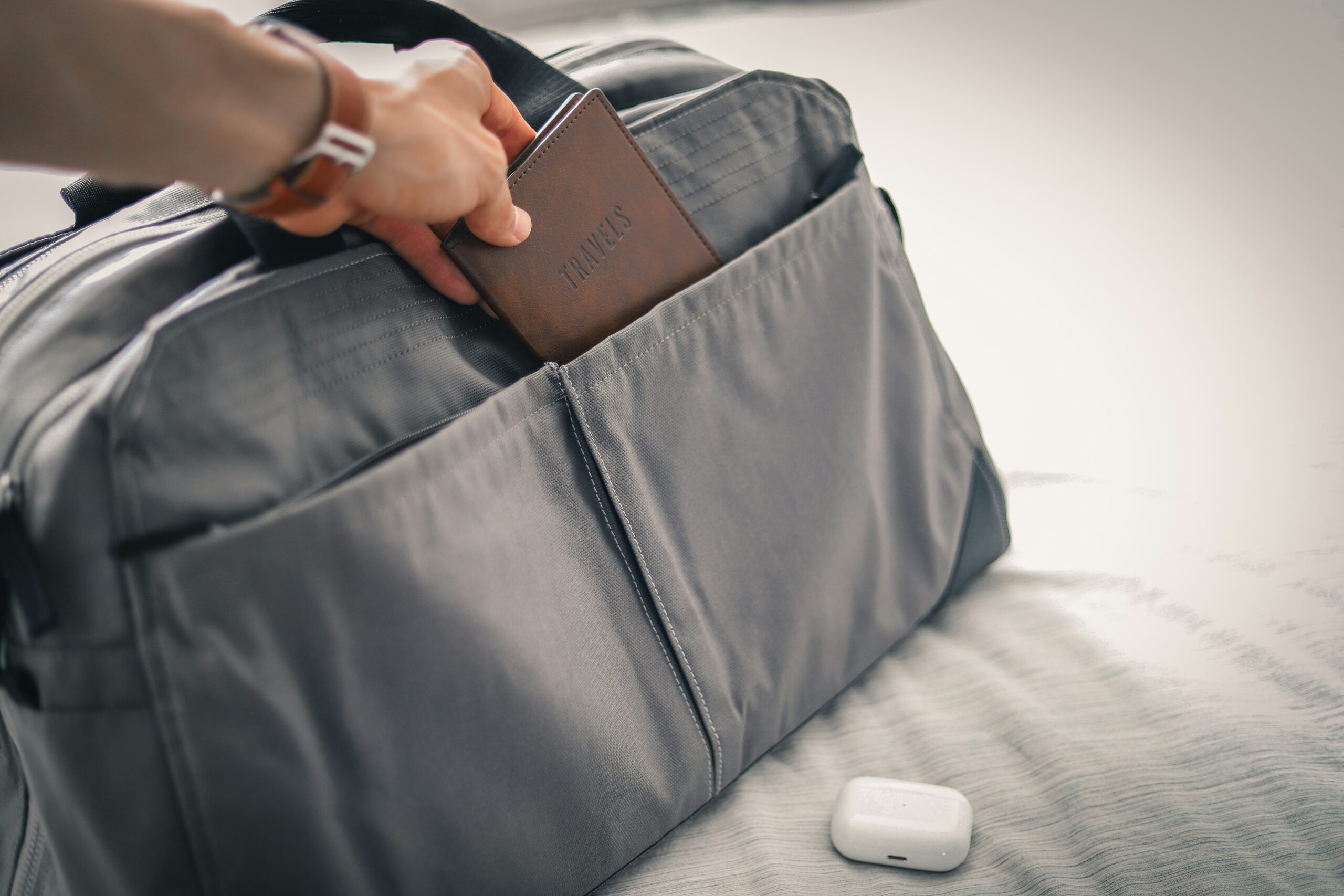 A person places a brown travel wallet labeled “TRAVELS” into the front pocket of a gray travel bag resting on a bed, with a pair of white wireless earbuds beside it.