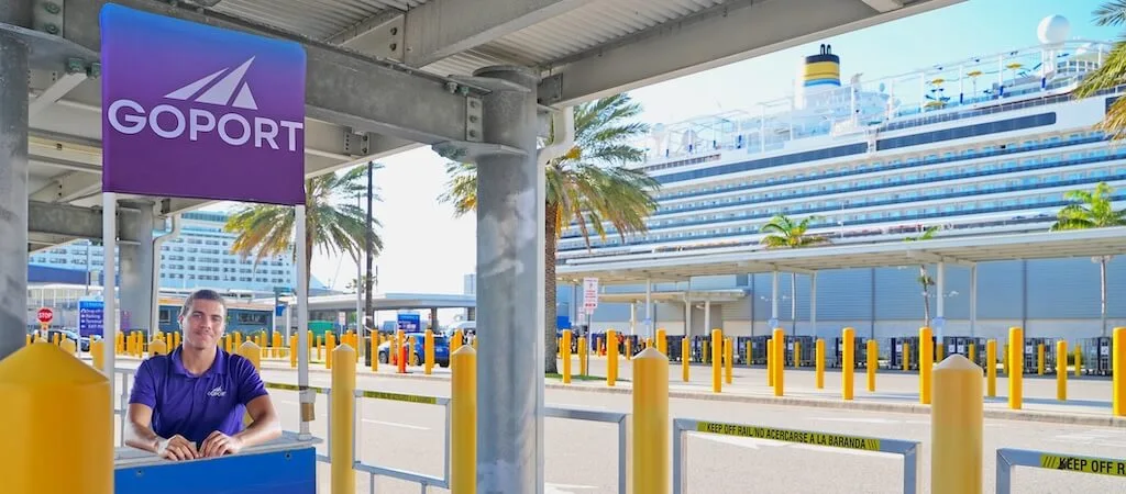 A Go Port staff member stands at a branded check-in podium at Port Canaveral with a cruise ship docked in the background.