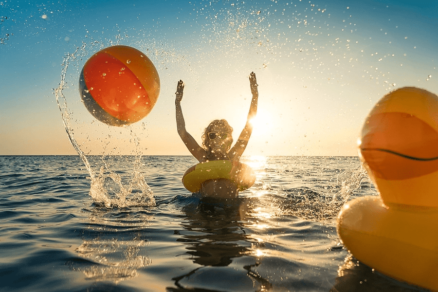 Woman in ocean throwing beach ball with sunset behind her