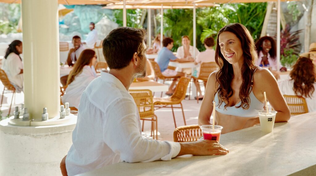 A couple enjoys tropical drinks together at an open-air bar surrounded by other guests and lush greenery.
