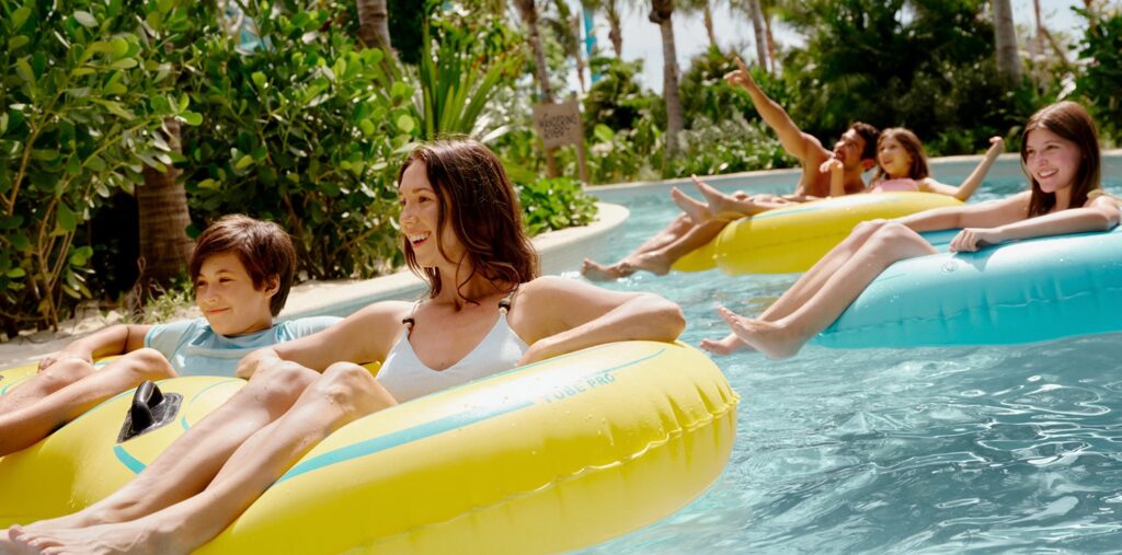 Families relax on colorful inner tubes while floating along a tropical lazy river surrounded by lush greenery.