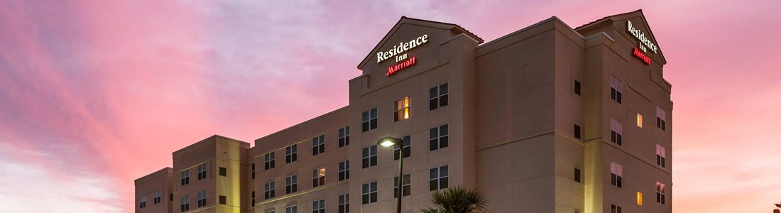 Exterior view of the Residence Inn by Marriott at sunset, featuring a multi-story building with illuminated windows and signage, under a pink and purple sky.